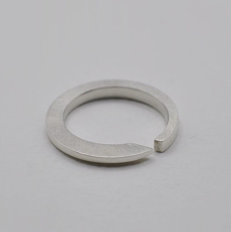 Querschnittsverformter Ring mit schräger Öffnung aus Silber liegt auf weißem untergrund