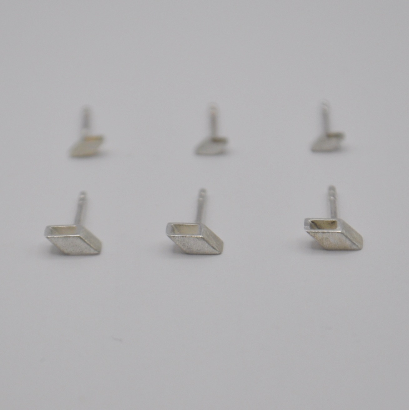 Sechs Ohrstecker in schräger, quadratischer Form. Drei größere im Vordergrund, kleine im Hintergrund
