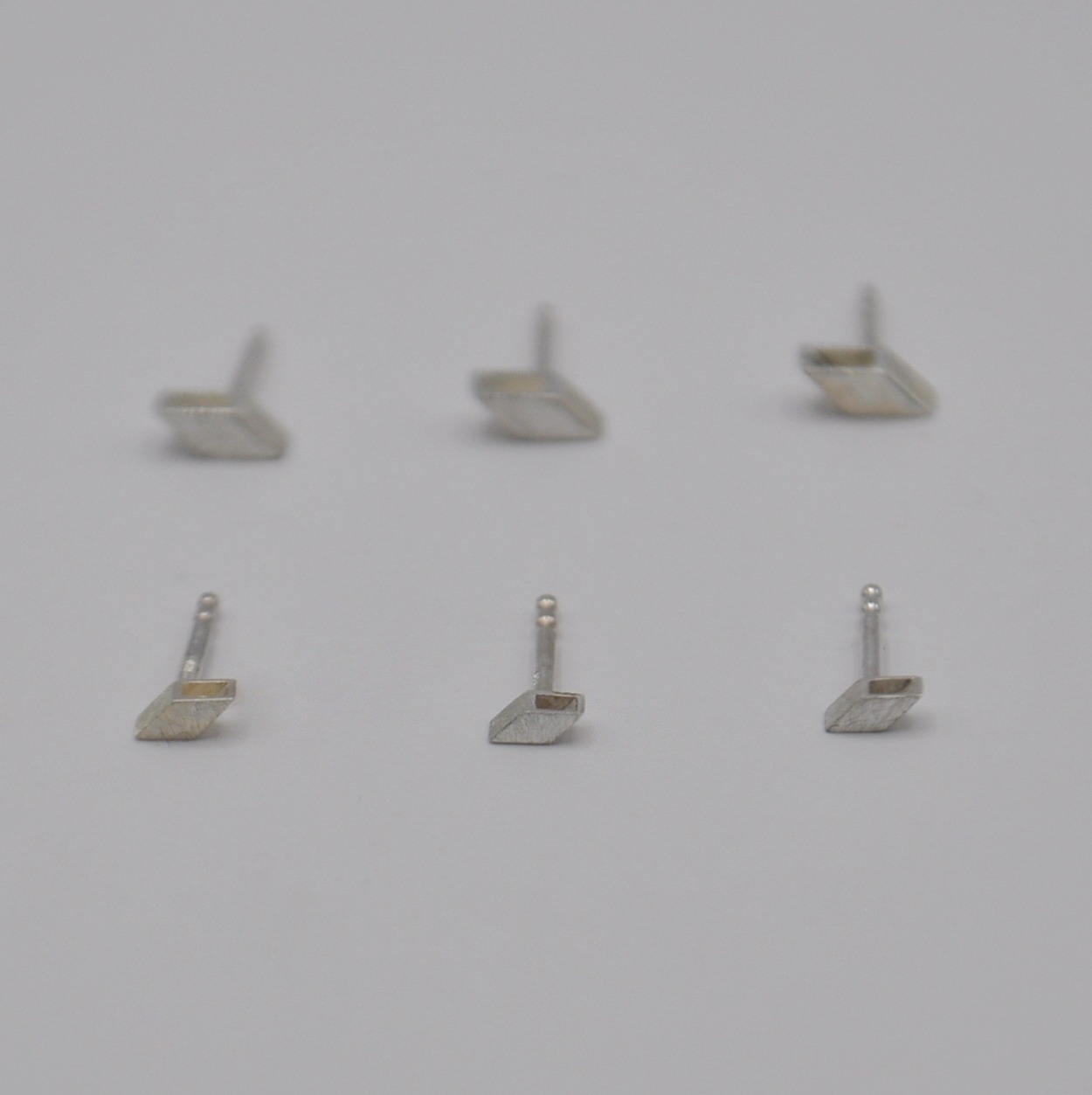 Sechs Ohrstecker in schräger, quadratischer Form. Drei kleine im Vordergrund, größere im Hintergrund