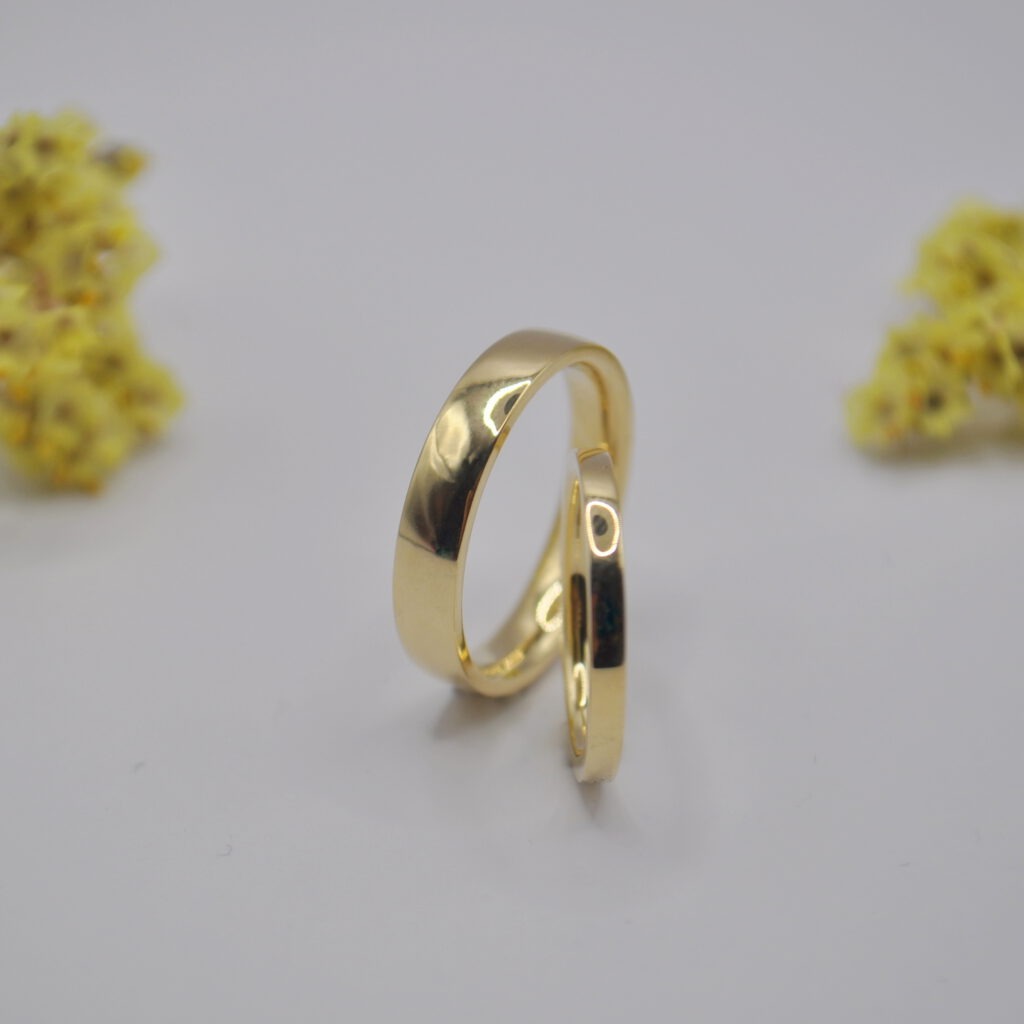 Paar gelbgoldene Trauringe mit gradlinigem Profil steht aufrecht auf hellem Untergrund